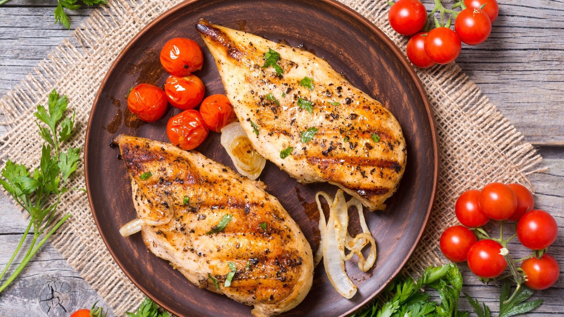 Two grilled chicken breasts on a black plate, accompanied by grilled tomatoes loading=
