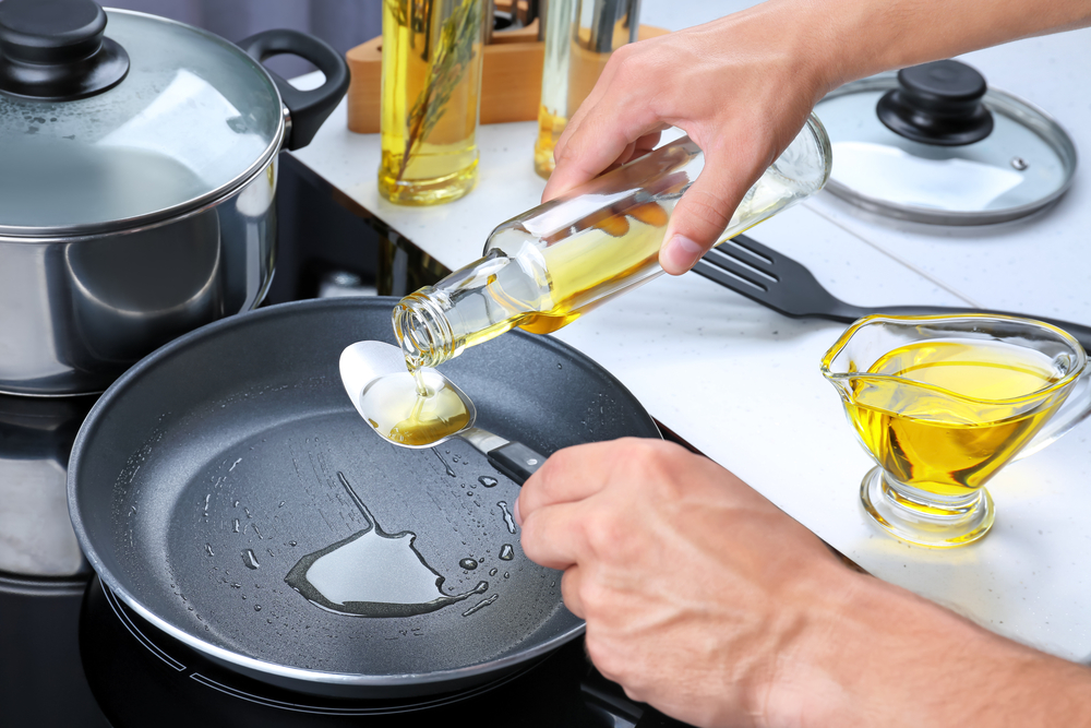A pair of elderly hands pouring oil into a frying pan, using a spoon to measure the correct amount. loading=